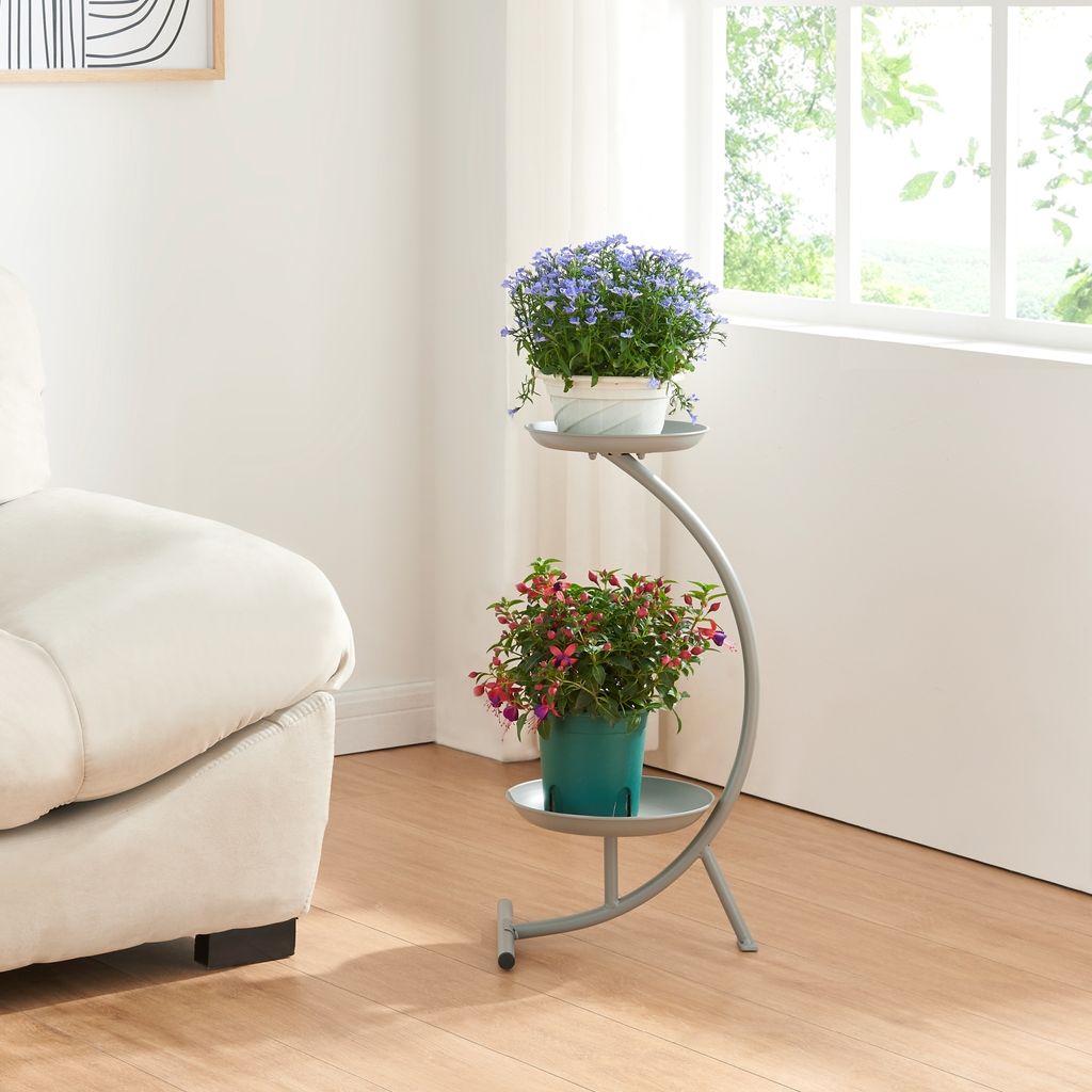 A plant stand with two potted plants next to a sofa and a window.
