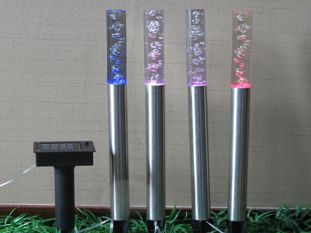 Four solar-powered garden lights with stainless steel bases and clear acrylic tubes filled with bubbles, lit in blue, pink, purple, and red. A solar panel unit is visible to the left.