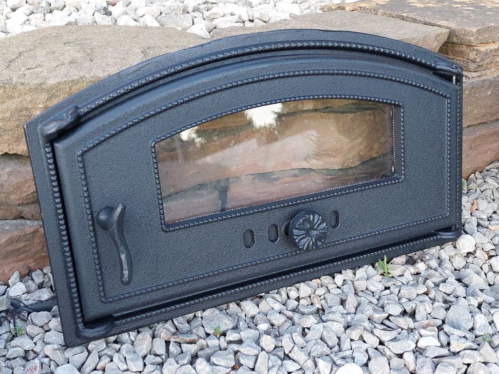 A black cast iron oven door with a glass window is placed on a bed of gravel and rocks.