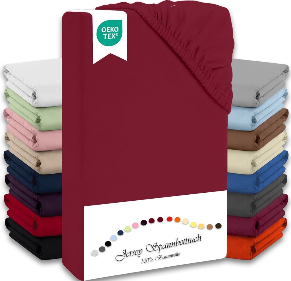 A stack of folded jersey fitted sheets in various colors, with a prominent bordeaux sheet in the foreground. A white label with a green OEKO-TEX logo is attached to the bordeaux sheet.