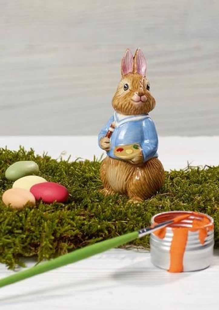 A ceramic bunny figurine dressed in a blue jacket, holding a palette and paintbrush, surrounded by moss and Easter eggs, with a can of orange paint and a paintbrush in the foreground.