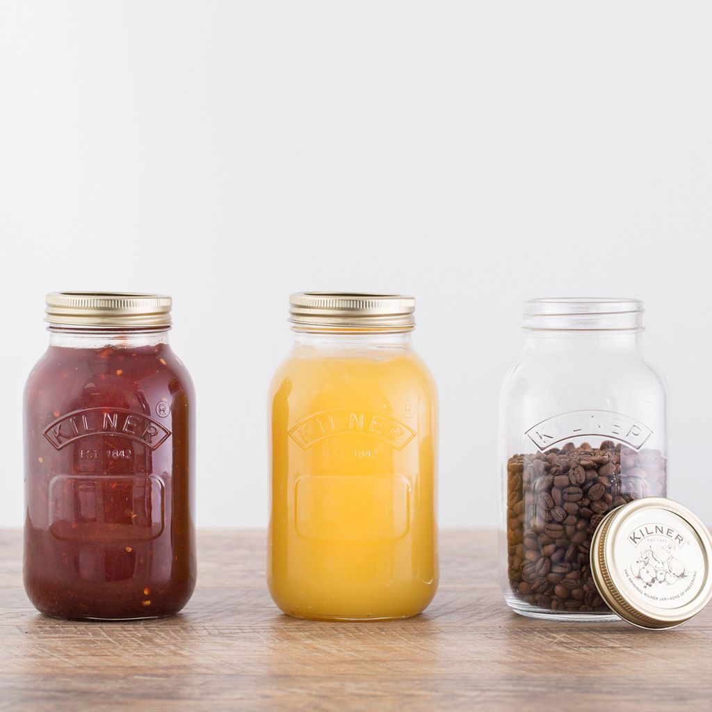 Three Kilner jars are arranged on a wooden surface against a white background. The leftmost jar contains a dark red substance, likely jam or sauce. The middle jar holds a bright yellow liquid, possibly juice. The rightmost jar is empty and contains coffee 