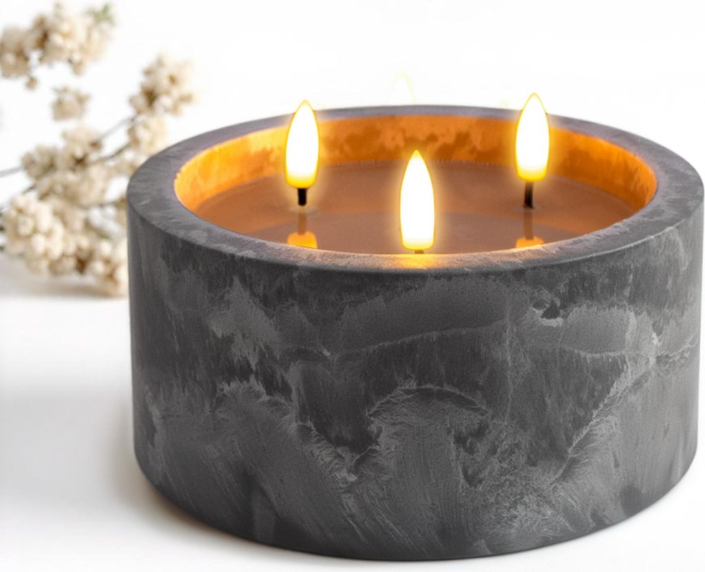 A three-wick LED candle in a concrete-look holder, with a sprig of white flowers in the background.