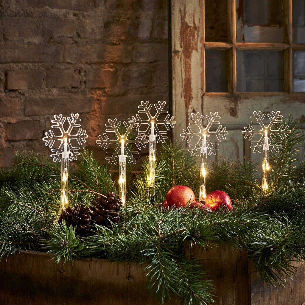 Four snowflake-shaped LED lights are placed among pine branches, with two red apples and pinecones.