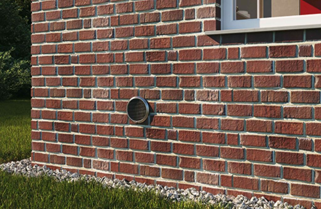 A vent on a red brick wall of a house, with grass and gravel at the base and a window sill above.