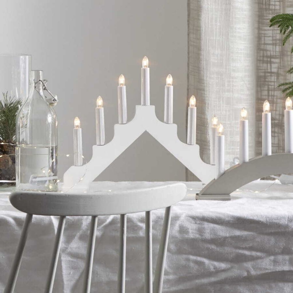 A white candle bridge light is displayed on a table covered with a white tablecloth, with a glass bottle and greenery in the background.