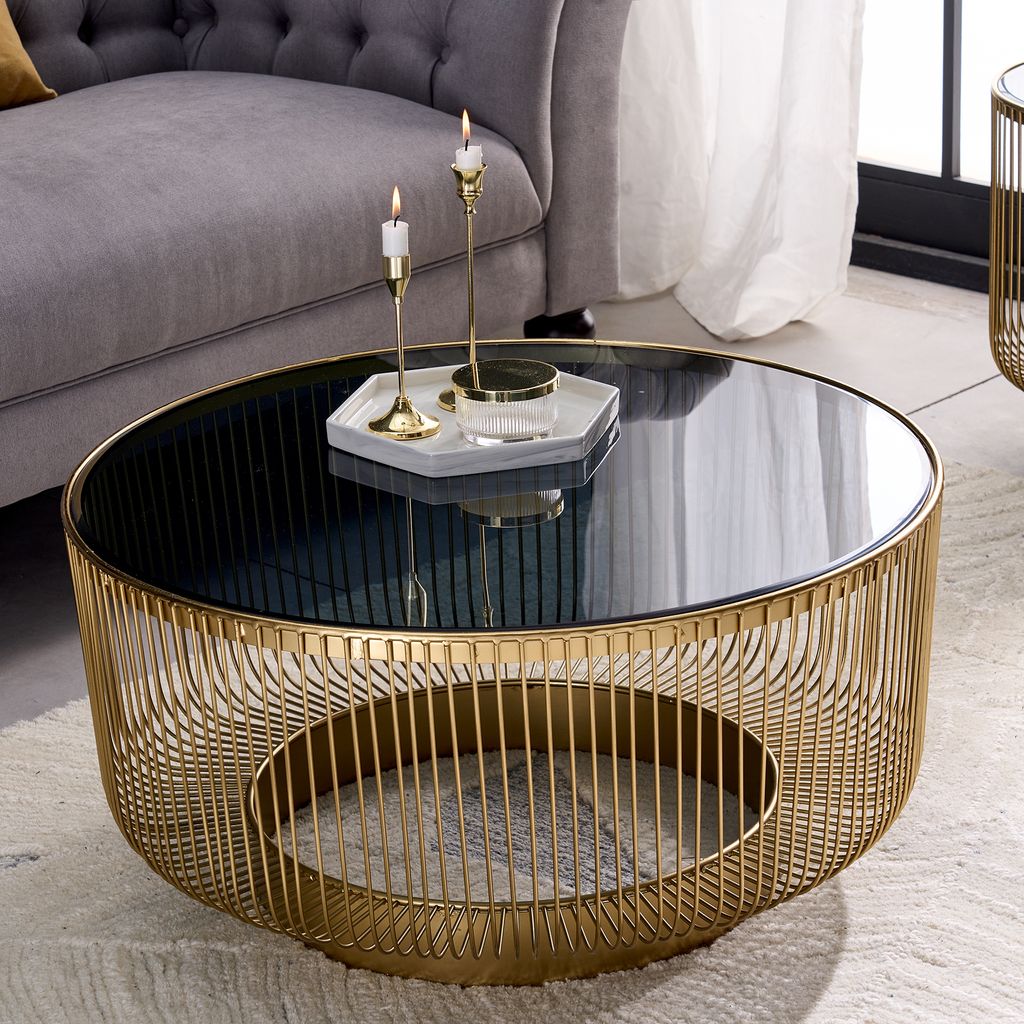 A round coffee table with a gold metal frame and a black glass top, placed in a living room setting next to a gray sofa.