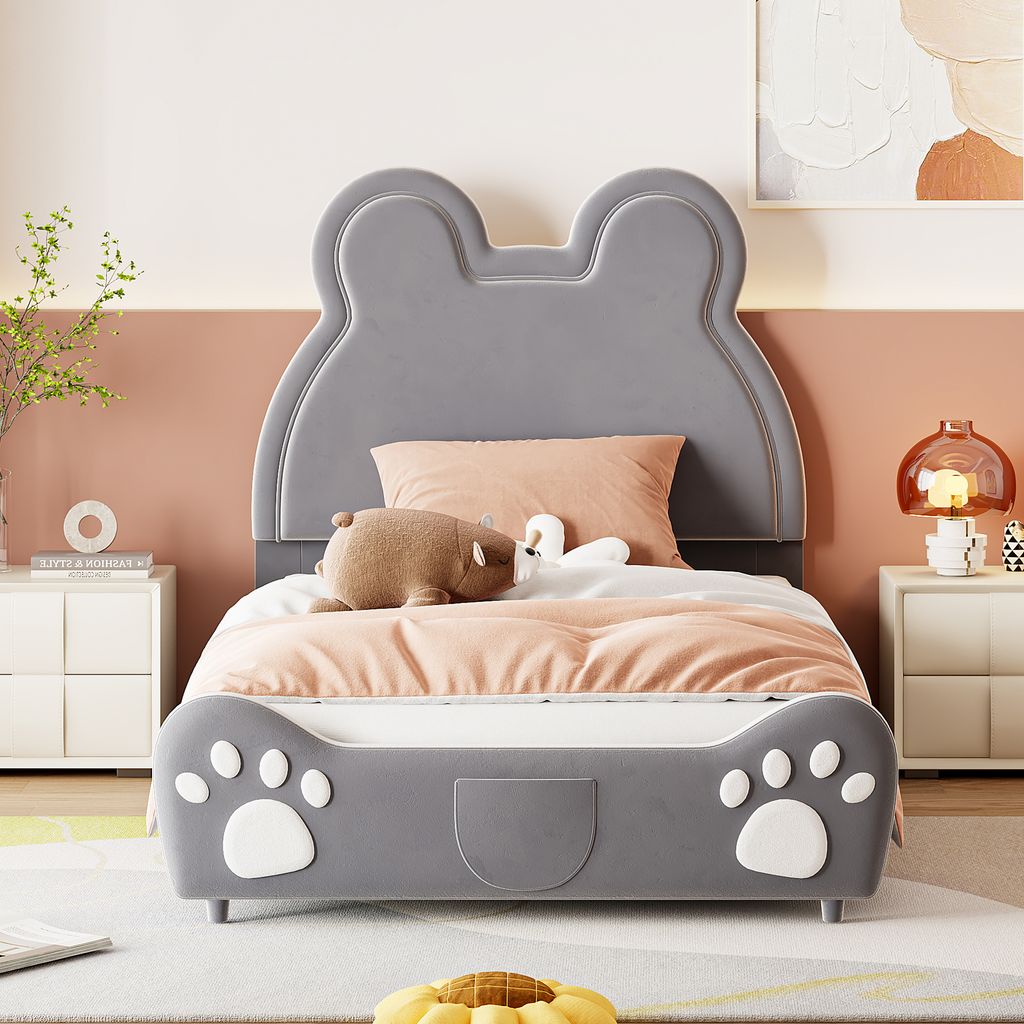 A child's bed with a bear-ear headboard and paw print details on the footboard, set in a bedroom with a pink and white wall, bedside tables, and a lamp.