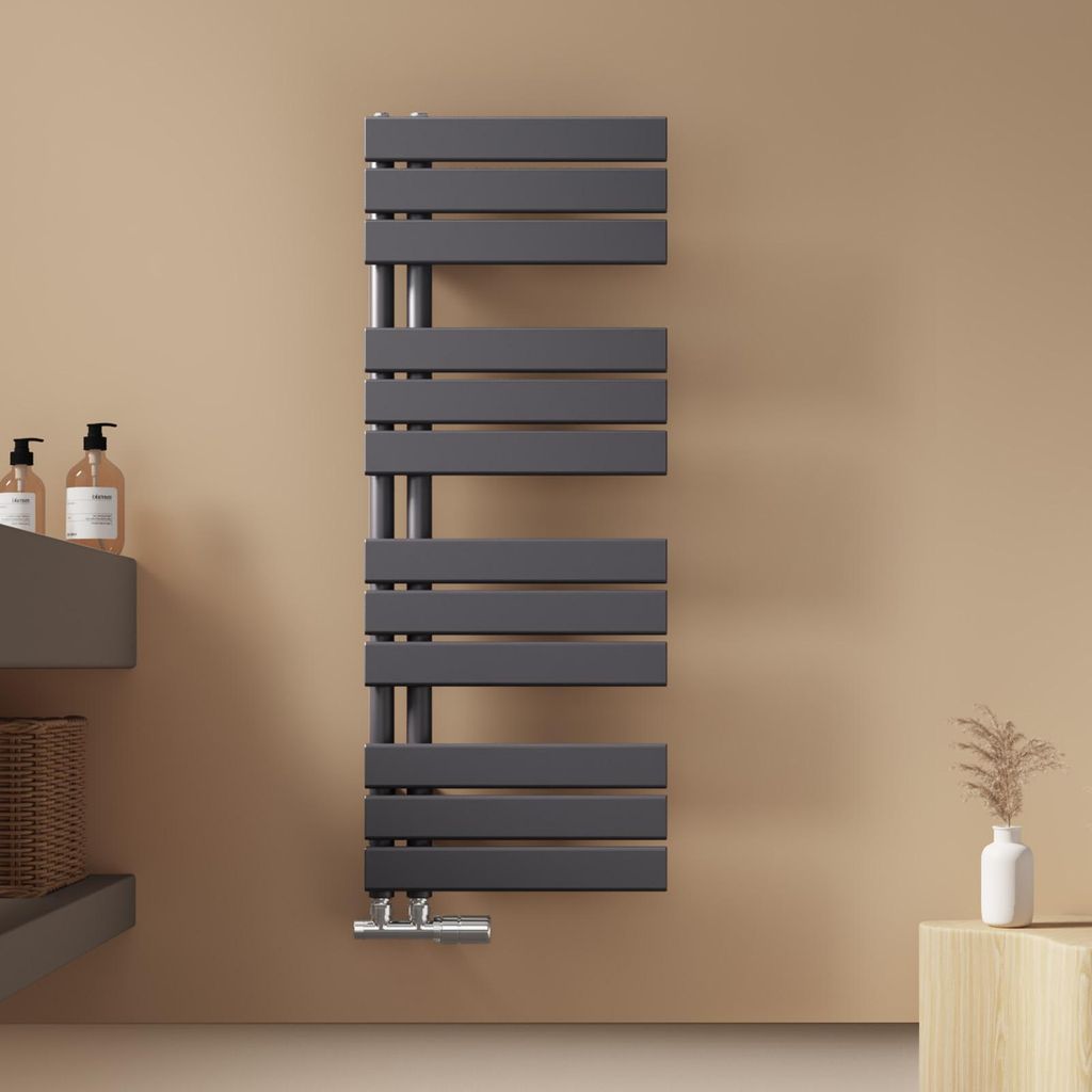 A dark gray towel radiator mounted on a light brown wall, with bathroom accessories on a shelf to the left and a small vase with dried grass on a wooden block to the right.