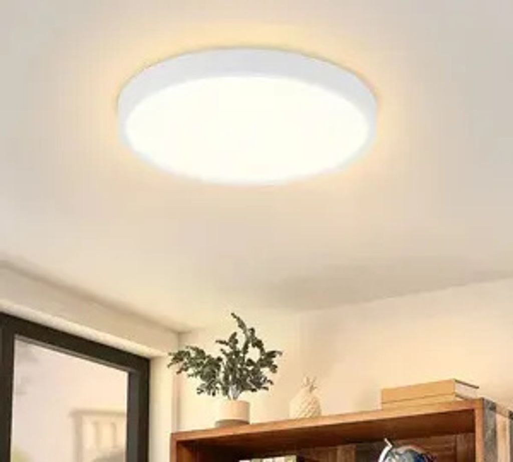 A white, circular LED ceiling light is mounted on a white ceiling. Below the light, a wooden shelf holds a potted plant and some books.