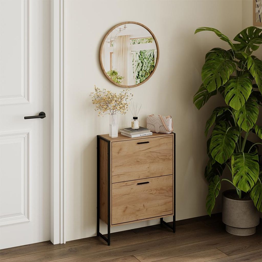 A shoe cabinet with two flip drawers is placed next to a door and a large potted plant. A round mirror hangs on the wall above the cabinet.