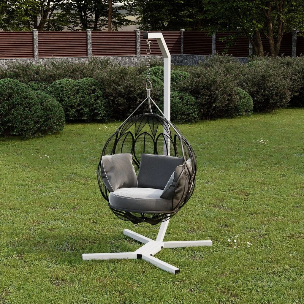 A hanging chair with cushions is placed on a grassy lawn in a garden setting, with bushes and a fence in the background.