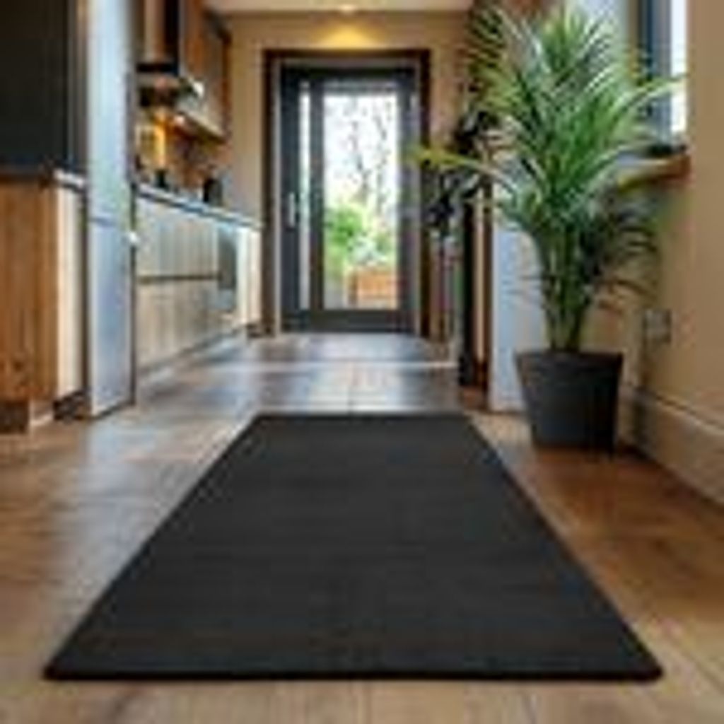 A dark rug is placed on a wooden floor in a hallway, with a potted plant and kitchen cabinets visible.