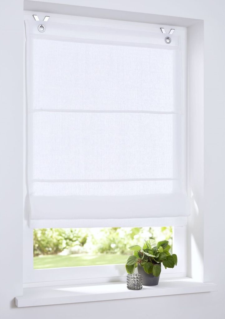 A white Roman blind is shown hanging in a window, with a view of greenery outside.
