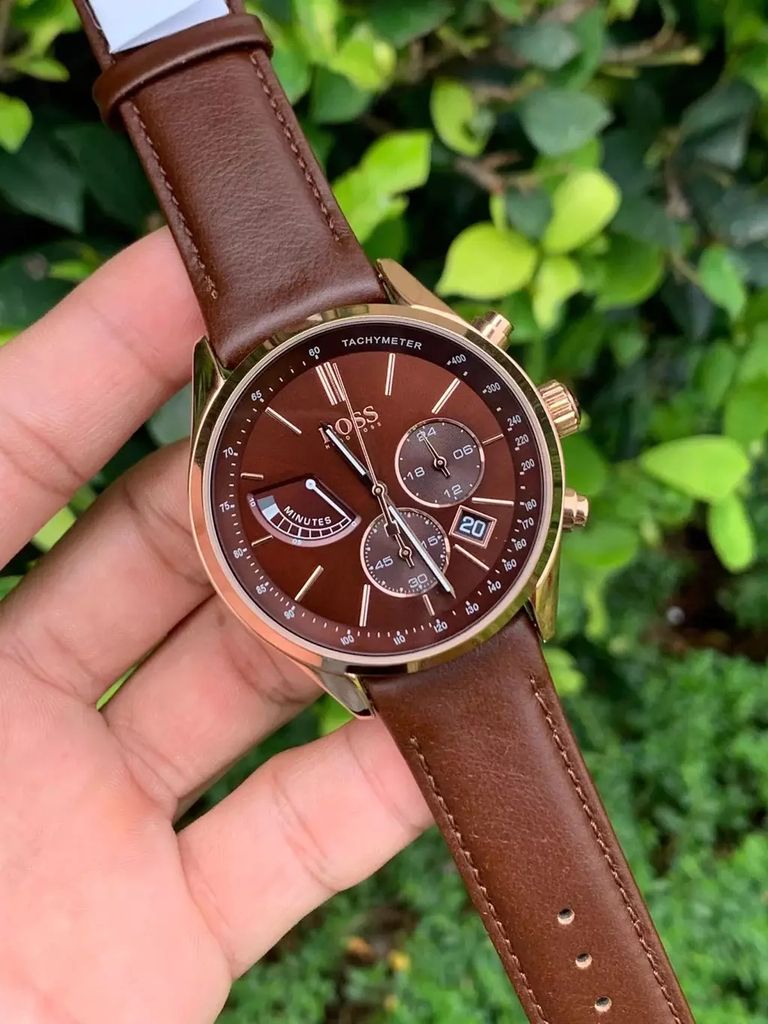 A hand holding a watch with a brown dial and strap, with a green leafy background.