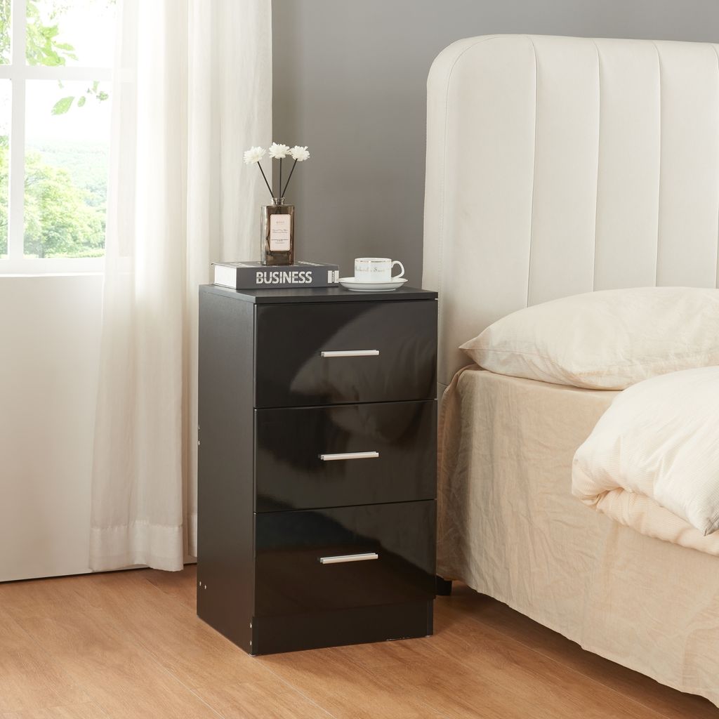 A black, high-gloss bedside table with three drawers is placed next to a bed with a white headboard and beige bedding. A window with white curtains is visible to the left.
