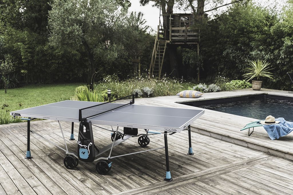 An outdoor ping pong table is set up on a wooden deck next to a swimming pool.