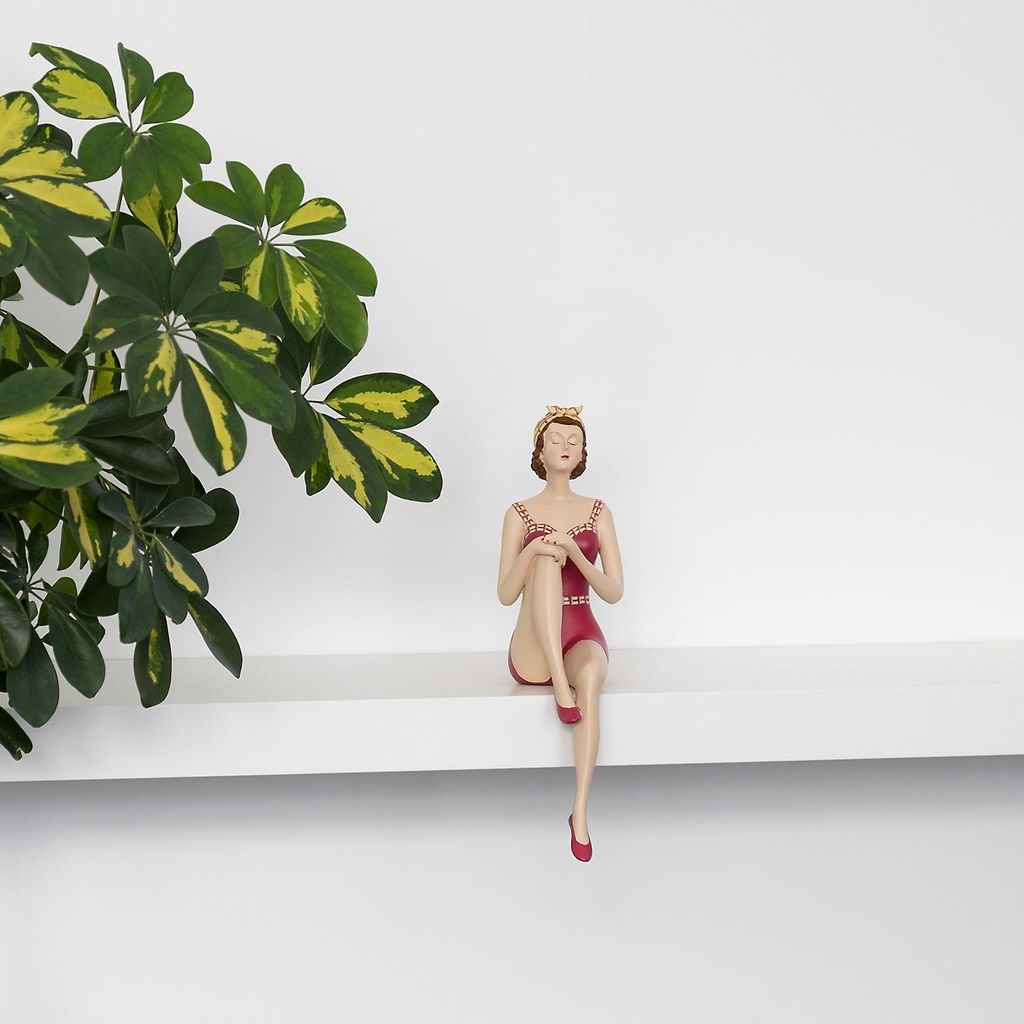 A decorative figurine of a woman in a red swimsuit sitting on a shelf next to a plant.