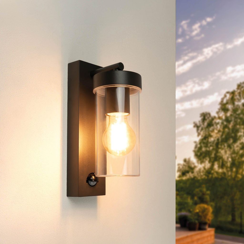A black outdoor wall light with a clear glass cylinder and a visible light bulb is mounted on a white wall. The background shows a blurred view of trees and sky.