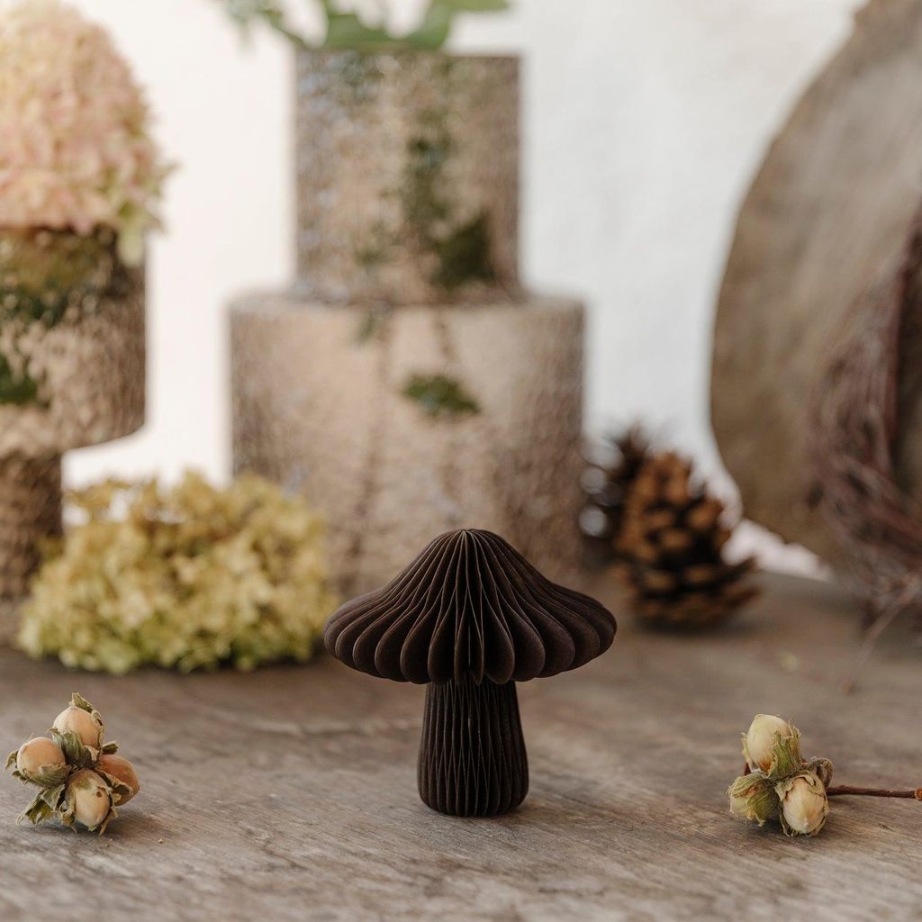 A dark brown paper mushroom decoration sits on a wooden surface, surrounded by natural elements like hazelnuts and dried flowers.