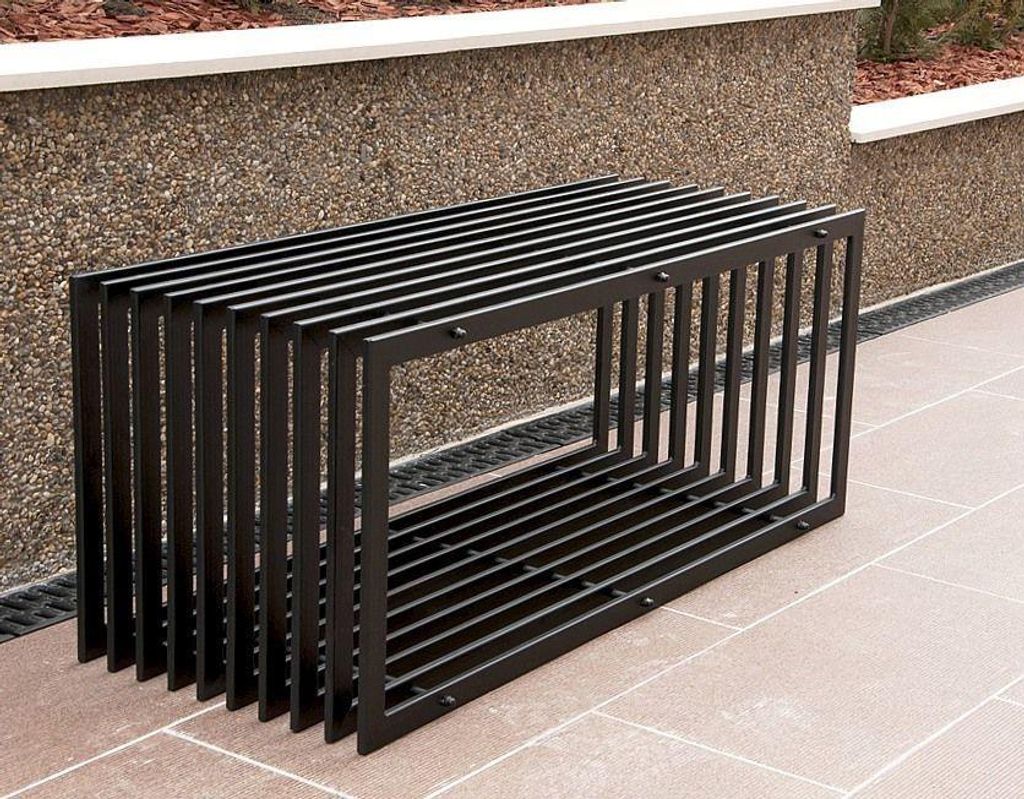 A modern black metal garden bench is placed outdoors on a paved patio, with a textured stone wall and landscaping in the background.