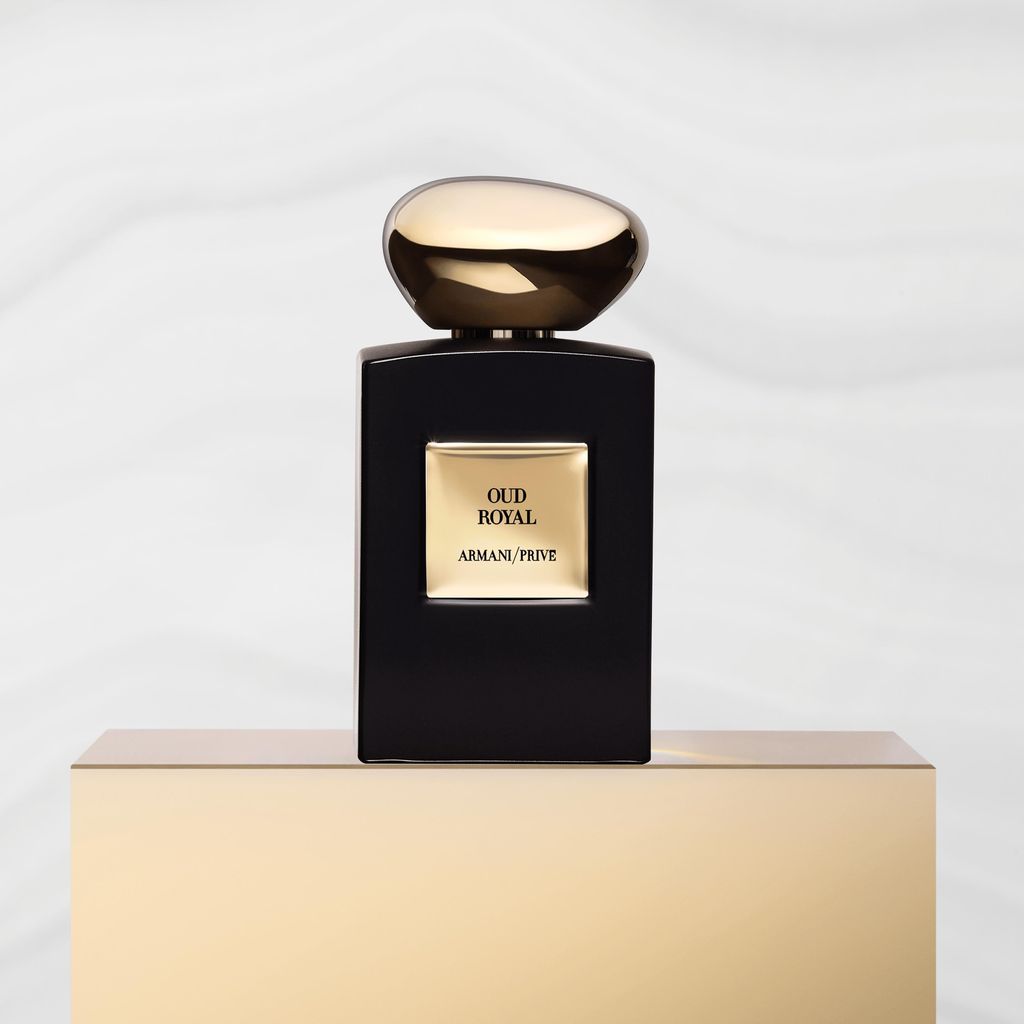 A black perfume bottle with a gold cap and a gold label sits on a gold pedestal against a white and gray marbled background.