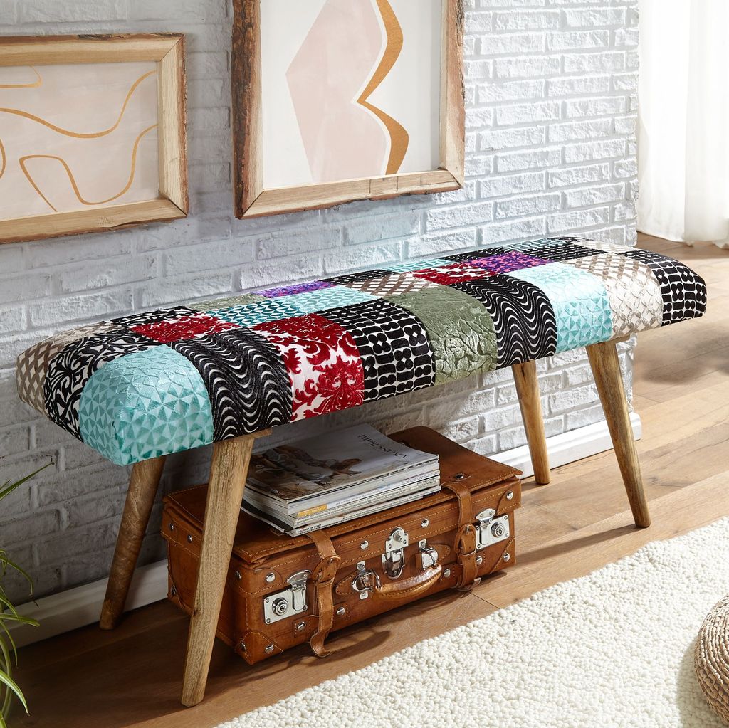 A patchwork bench is placed in a room with a brick wall and artwork, with a vintage suitcase and magazines underneath.