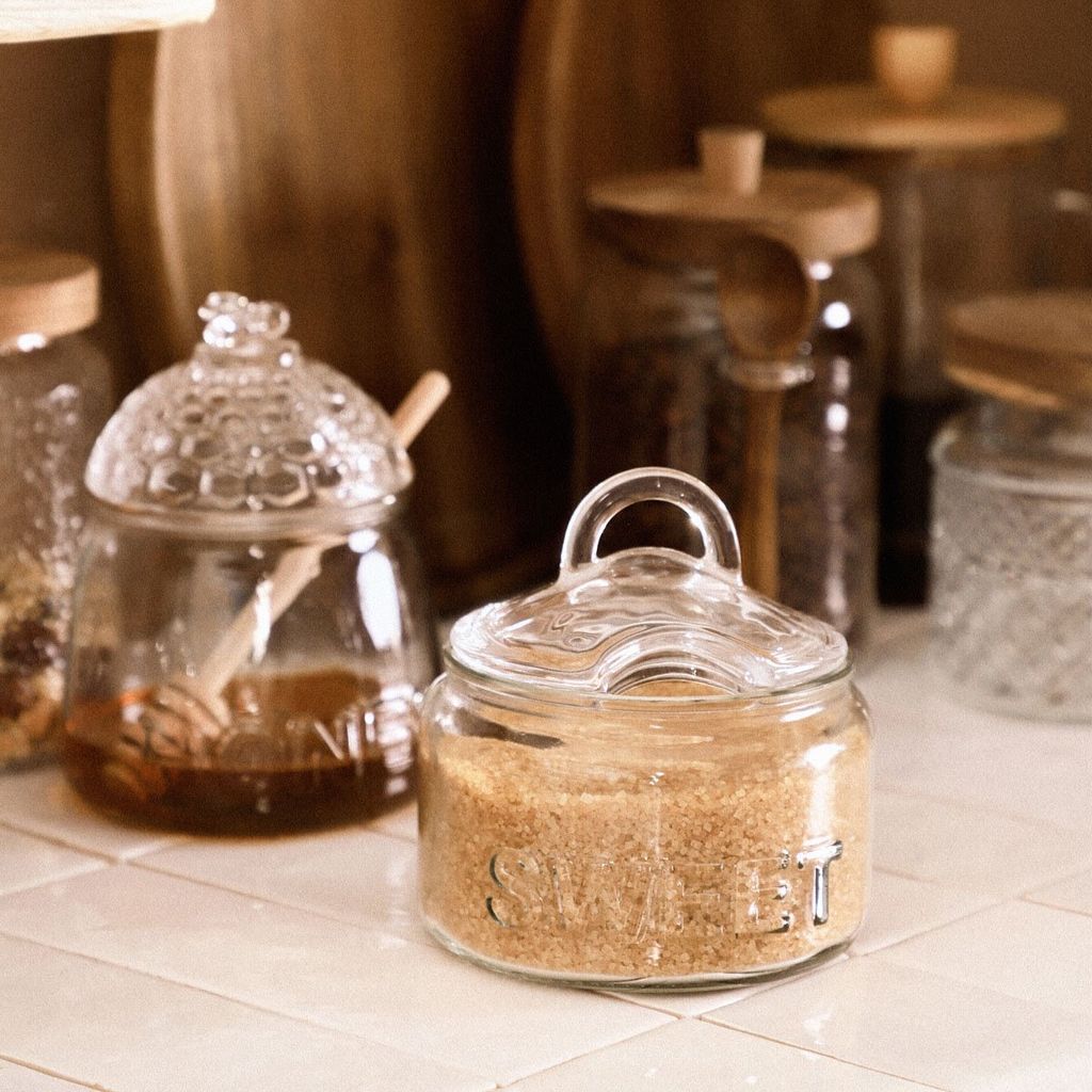 Two glass jars with lids, one containing honey and a honey dipper, and the other containing brown sugar. They are placed on a tiled surface.