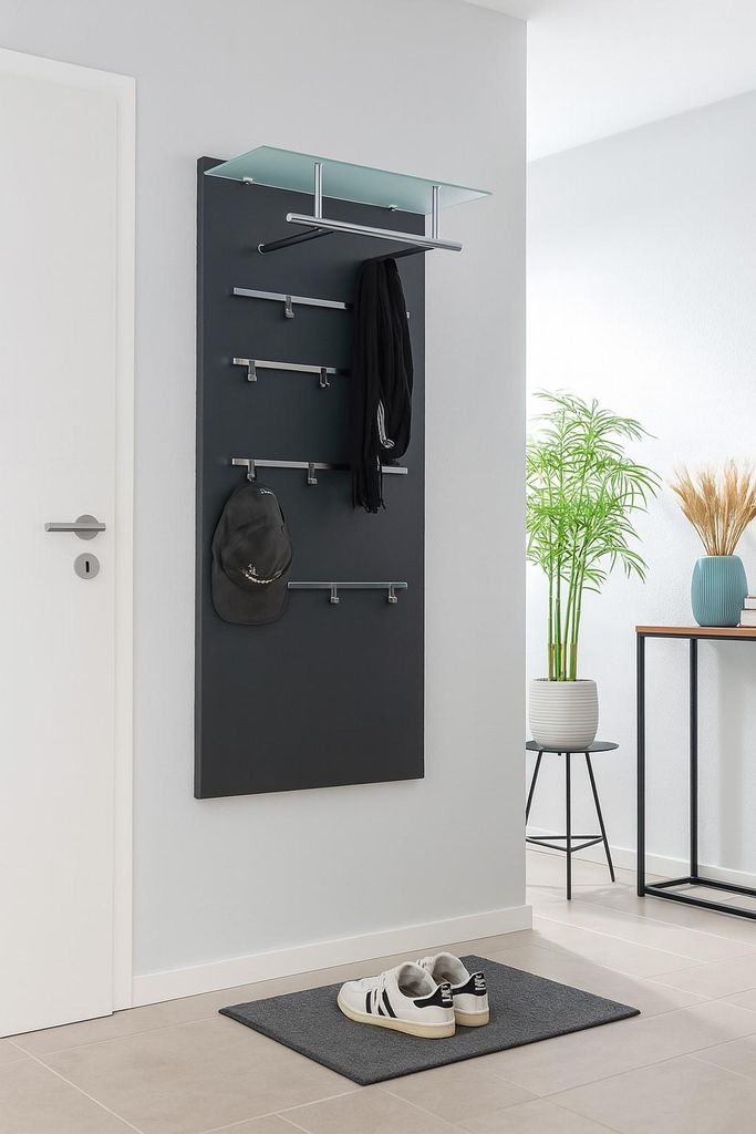 A modern entryway with a black coat rack panel mounted on a light gray wall, featuring hooks for hats and scarves, a glass shelf, and a pair of sneakers on a doormat.