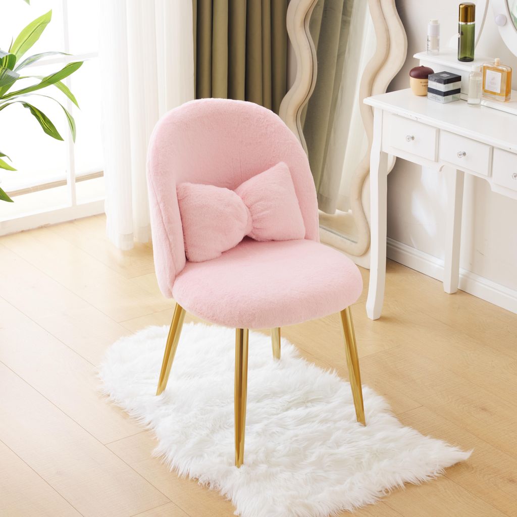 A pink fluffy chair with a bow cushion and gold legs sits on a white rug in front of a vanity table.