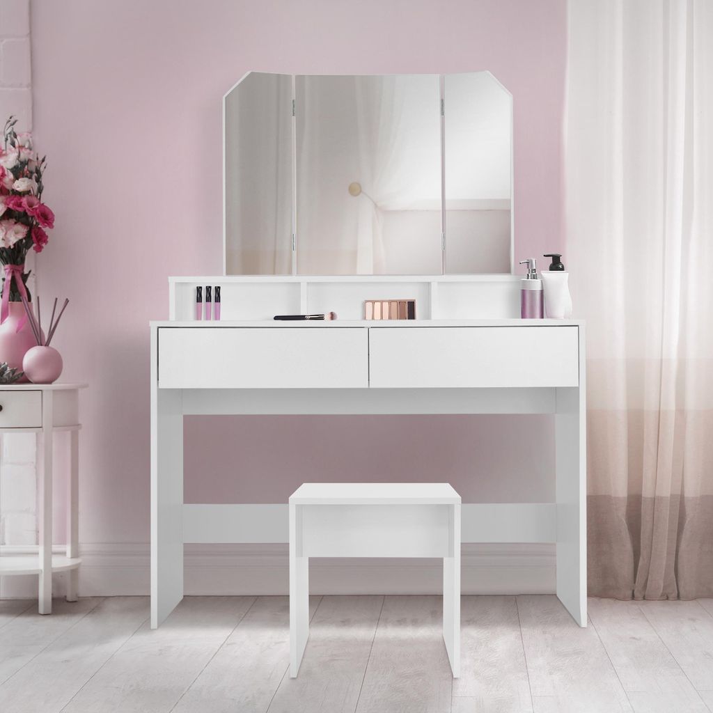 A white vanity table with a tri-fold mirror and a matching stool, set against a pink wall with sheer curtains in the background.