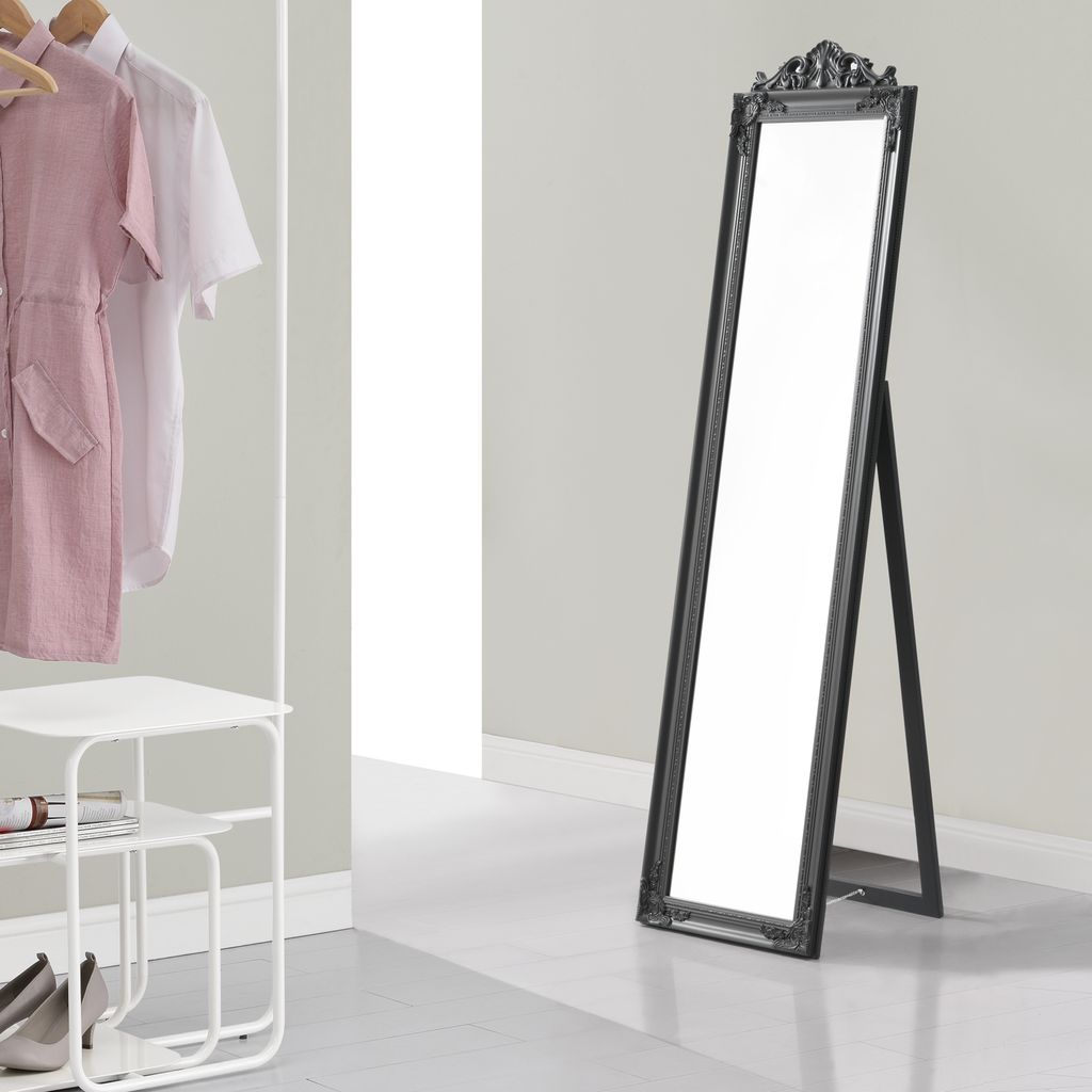 A full-length mirror with a dark gray frame stands on a light gray floor in a room with light gray walls. To the left, a white shelf unit holds shoes and a book, and a clothing rack displays a pink dress and a white shirt.