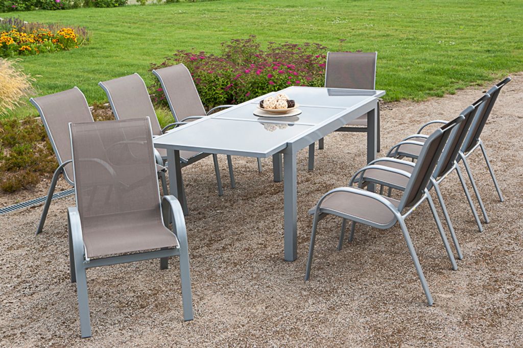 An outdoor dining set with a table and chairs on a gravel surface with a lawn and flowerbed in the background.