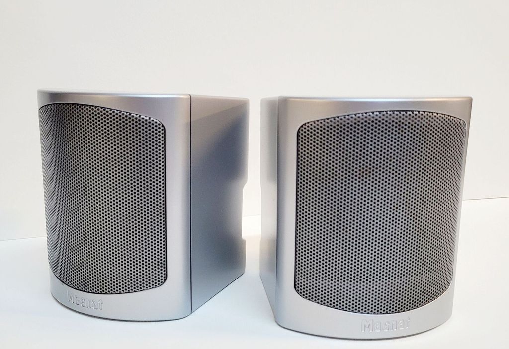 Two silver speakers are placed side-by-side on a white surface against a white background.