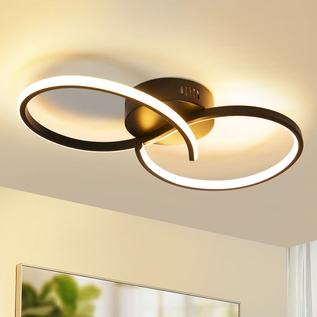 A modern black ceiling light with two intertwined rings, illuminated with warm white light, mounted on a white ceiling.