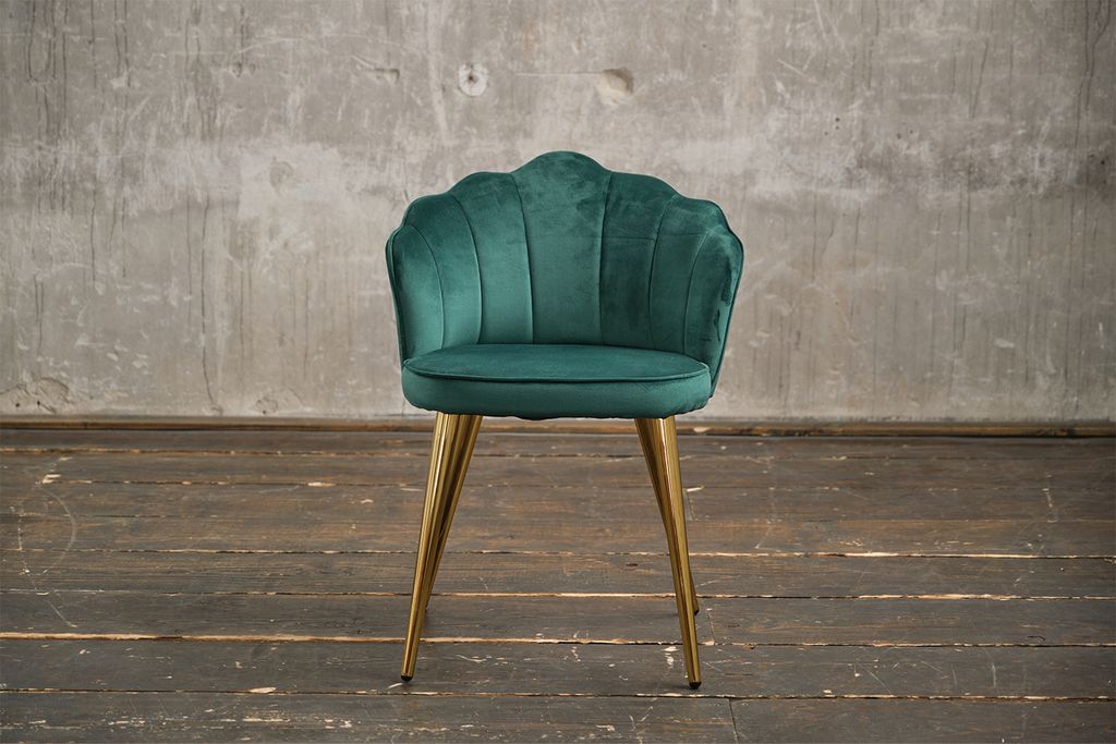 A green velvet chair with gold legs stands on a wooden floor in front of a concrete wall.