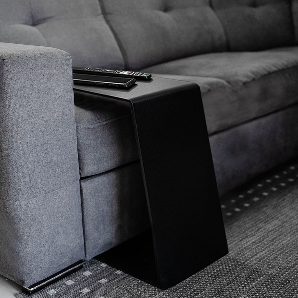 A close-up shot of a C-shaped side table placed next to a gray sofa, with two remote controls on top of the table.