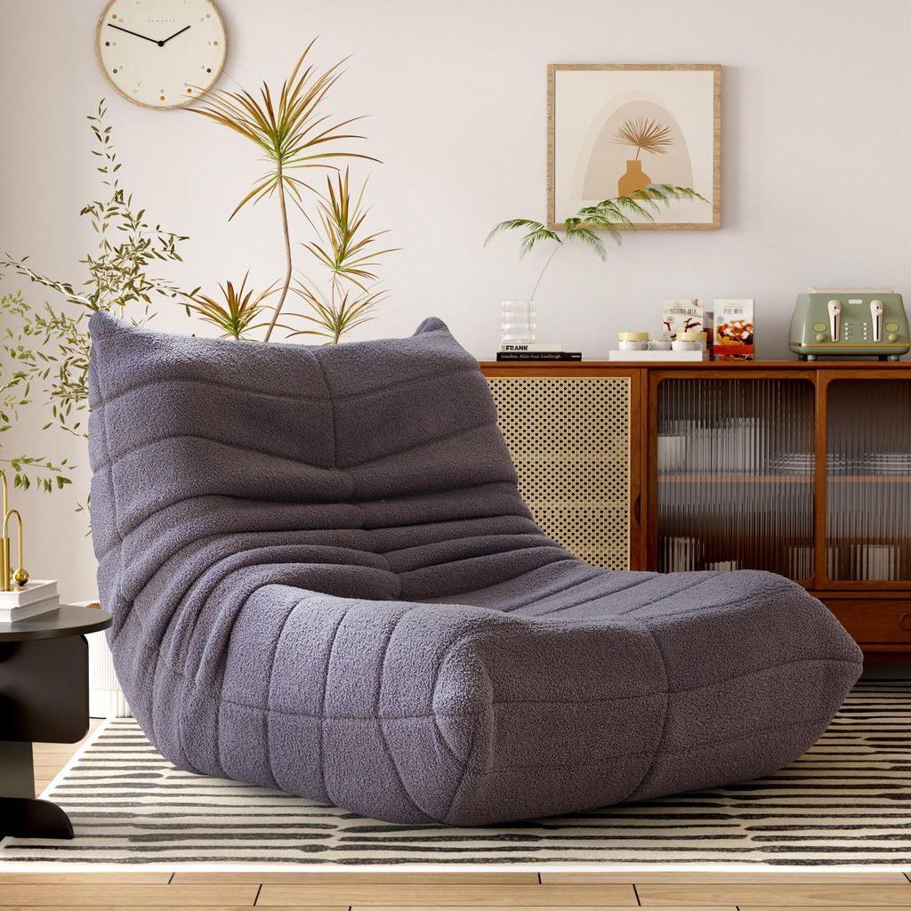 A gray beanbag chair is placed in a living room setting, next to a small black table and a rug. In the background, there is a wooden cabinet with glass doors, a clock on the wall, and a framed picture.