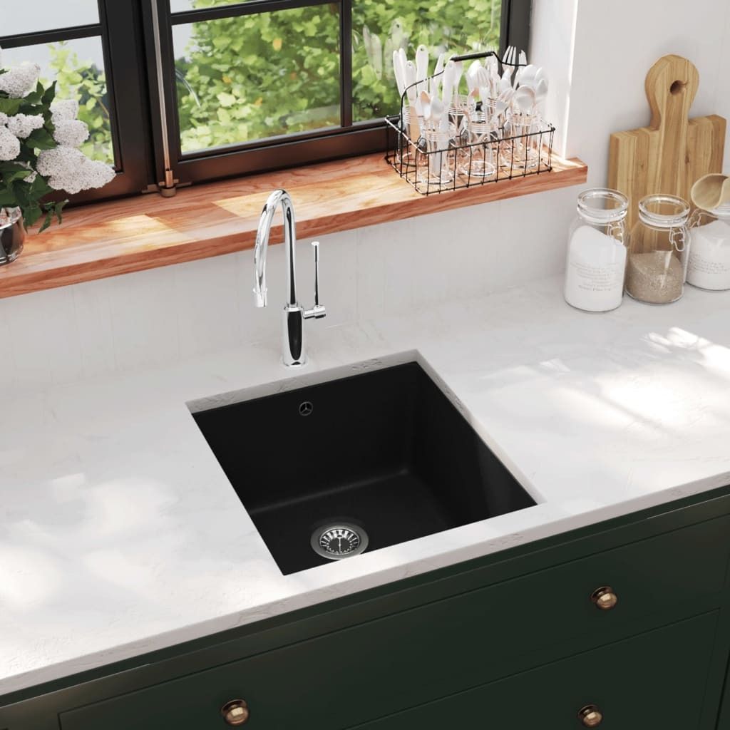 A modern kitchen sink with a black granite basin and a chrome faucet, set against a white countertop and dark green cabinets. A window with greenery is visible in the background.