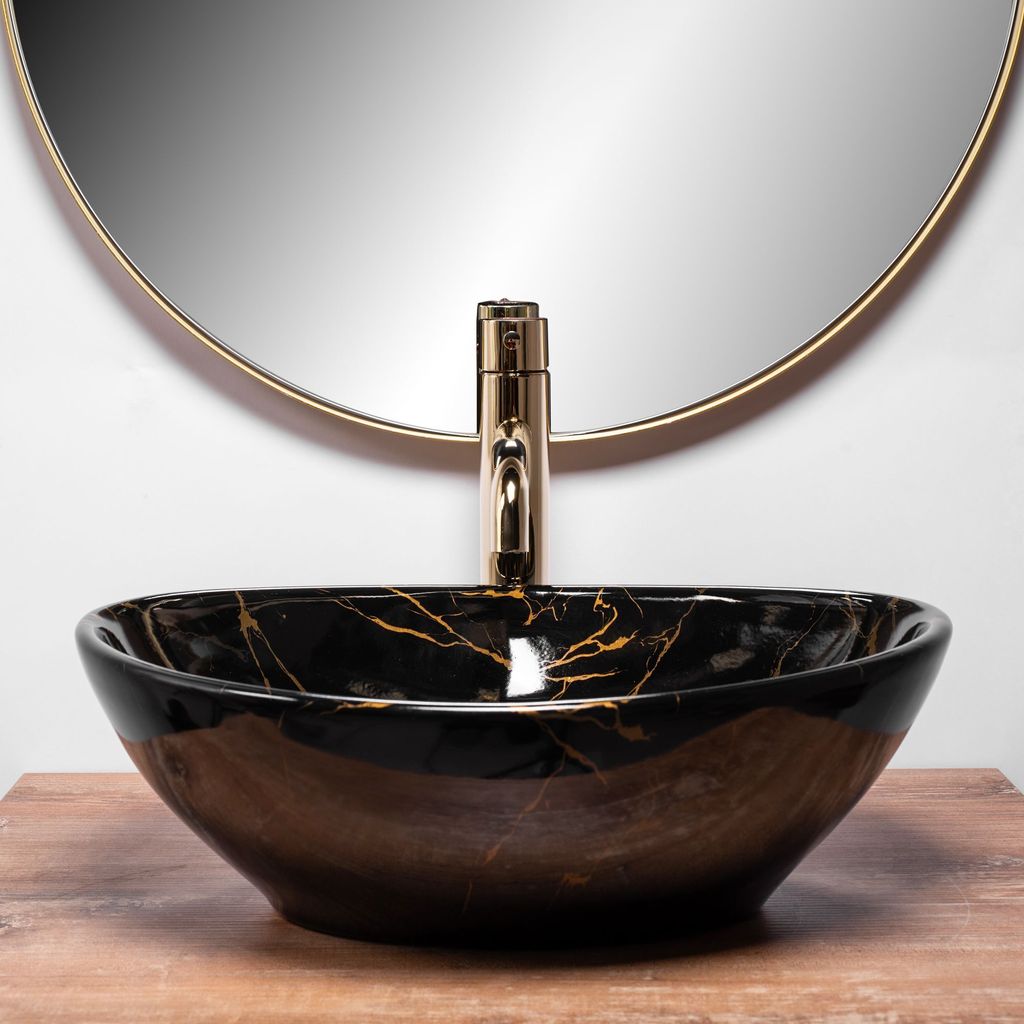 A black marble sink with gold veins sits on a wooden surface in front of a round mirror and a gold faucet.