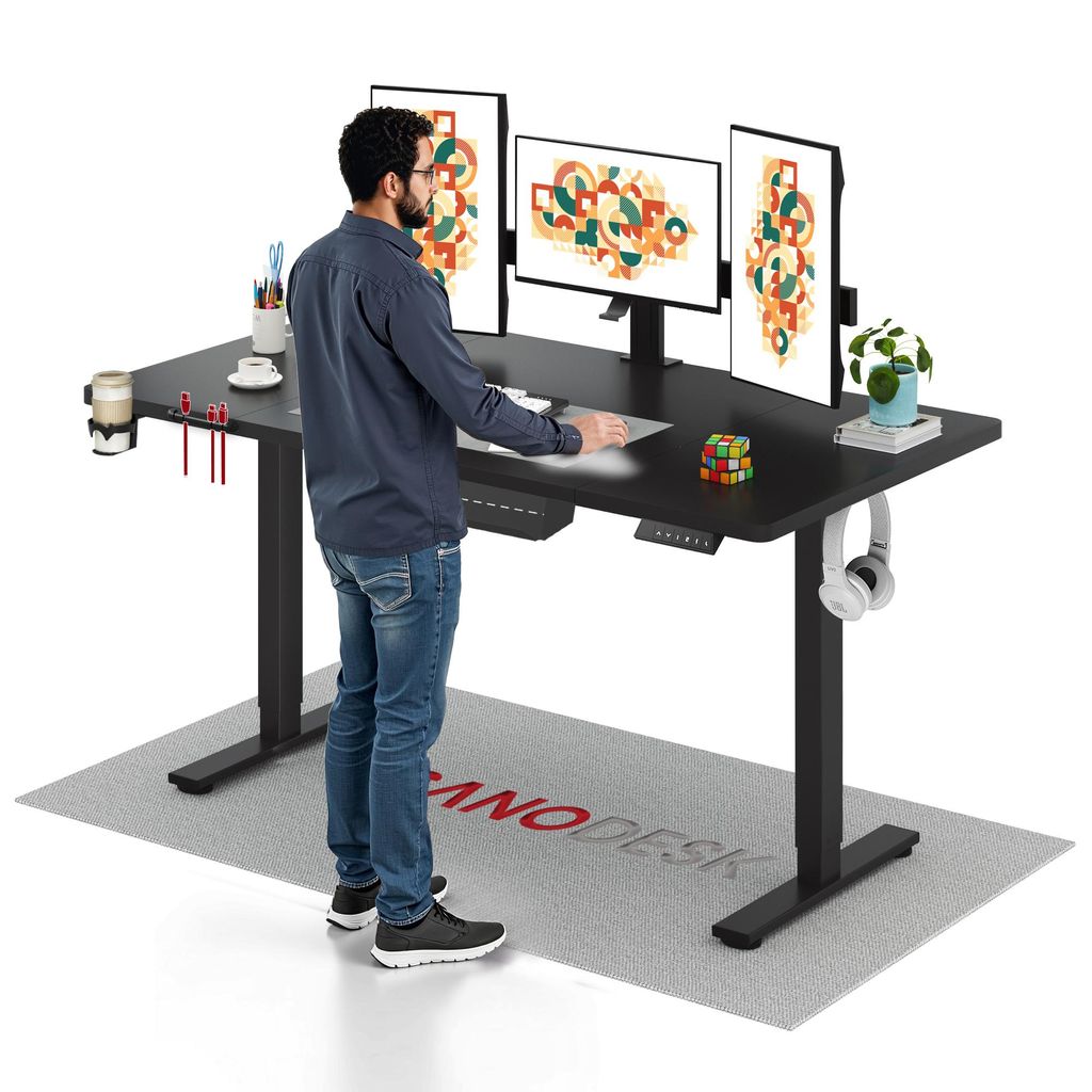 A man is standing at a black standing desk with three monitors, a keyboard, a mouse, and various desk accessories. A mat with a logo is on the floor in front of the desk.