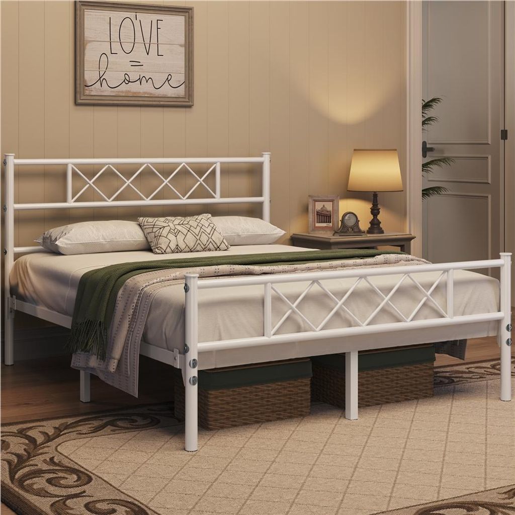 A bedroom scene featuring a white metal bed frame with a diamond pattern headboard and footboard, dressed with bedding and a green throw blanket. A bedside table with a lamp and decorative items is visible to the right.