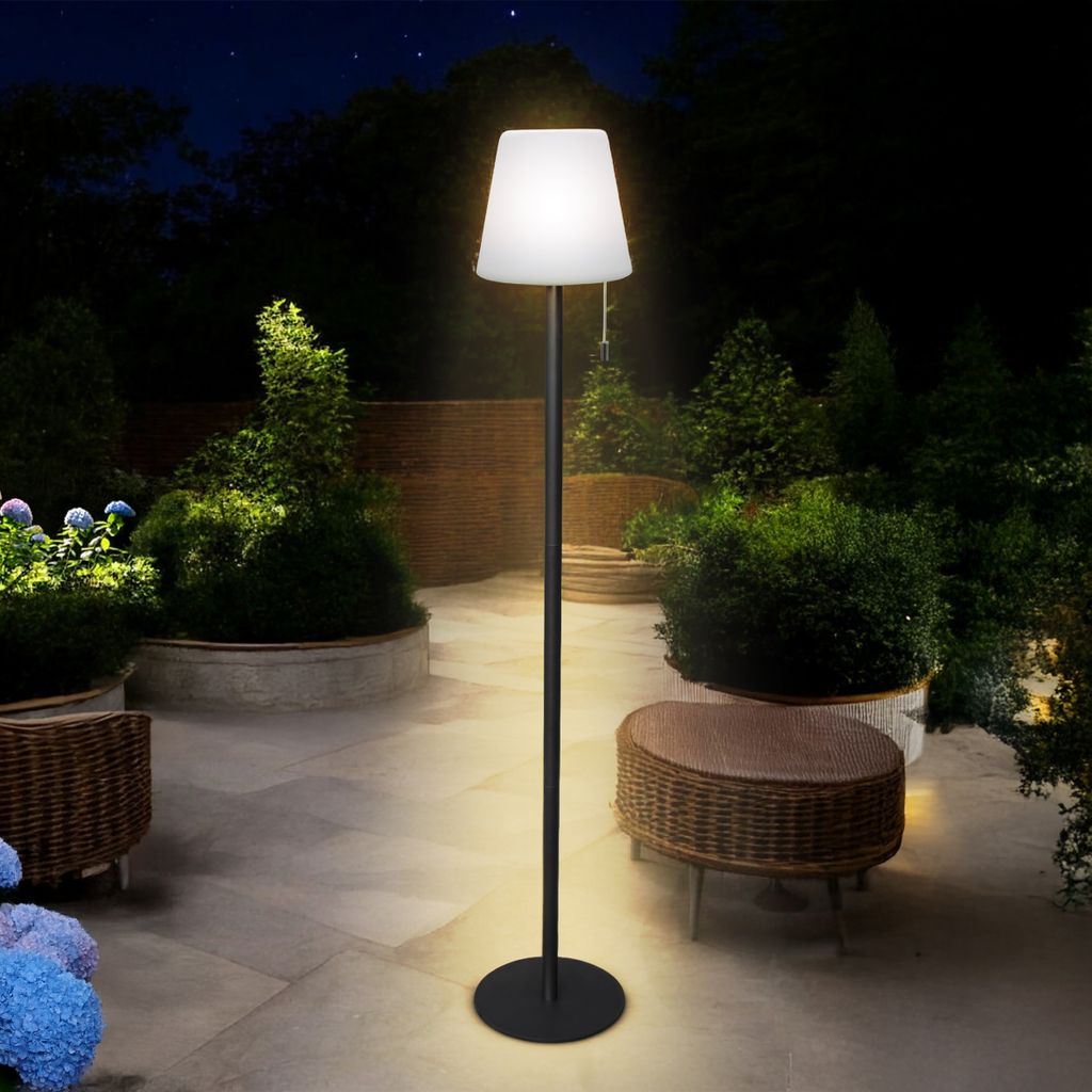 A modern floor lamp illuminates a patio at night, surrounded by lush greenery and outdoor furniture.