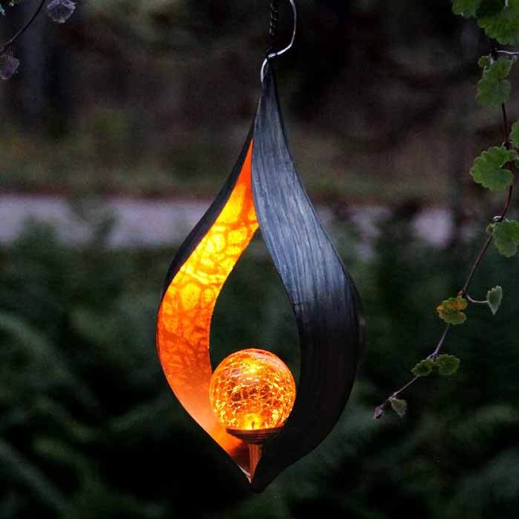 A decorative solar lantern hanging from a tree branch in a garden at dusk.