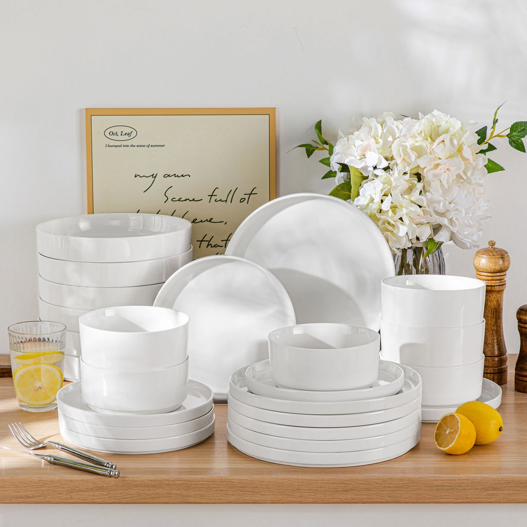 A collection of white ceramic dishes and bowls are arranged on a wooden surface, with a framed picture and a bouquet of white flowers in the background.