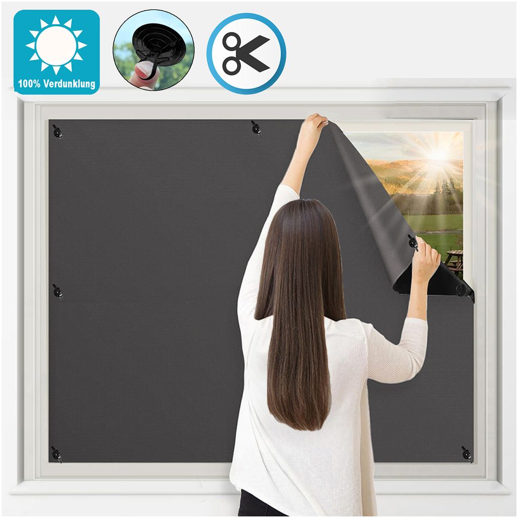 A woman is applying a dark gray blackout film to a window from the inside.