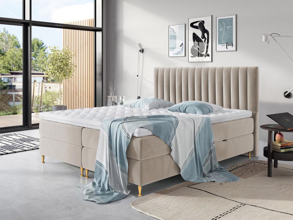 A bedroom scene with a large boxspring bed as the main focus, decorated with pillows and a throw blanket. Large windows with a view of greenery are visible in the background.