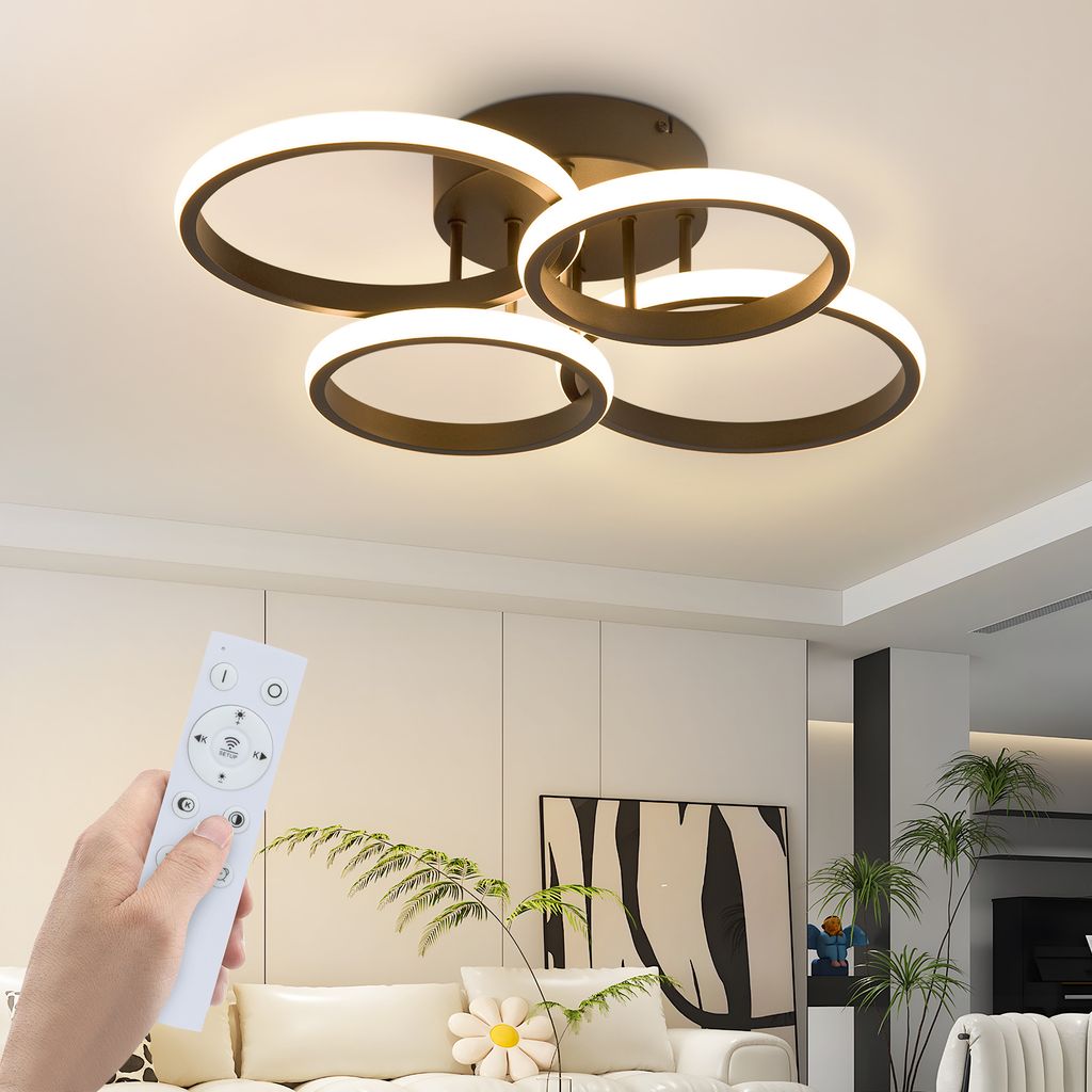 A hand holding a remote control in front of a modern ceiling lamp in a living room.