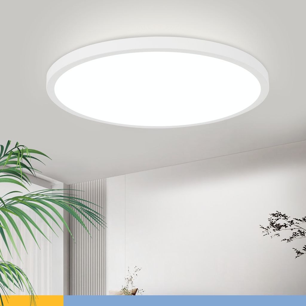 A round, white LED ceiling light is mounted on a white ceiling. A large green plant is in the foreground to the left, and a blurred background shows a wall with vertical paneling and a hint of another plant.