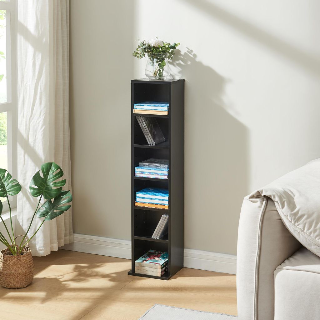 A tall, narrow black shelf unit filled with CDs and some books, placed in a bright room next to a large window with sheer curtains and a large potted plant.
